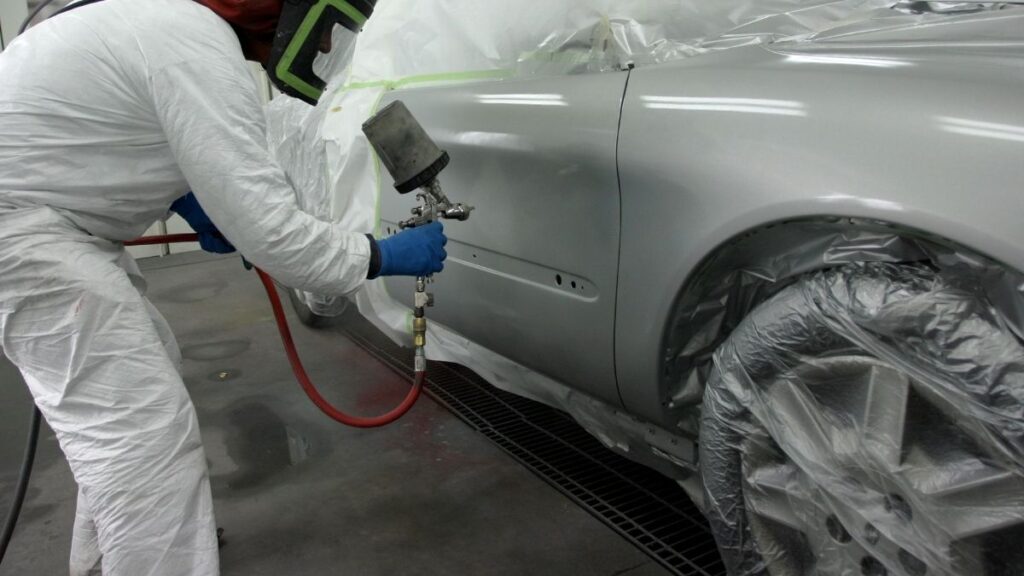 Pintor automotivo aplicando verniz em carro prata dentro de cabine de pintura